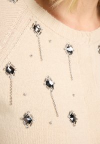 Beiger Pullover mit rundem Ausschnitt, versehen mit silbernen und schwarzen Verzierungen sowie verschiedenen geometrischen Mustern auf der Vorderseite.