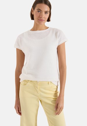 Femme aux cheveux courts marron porte un haut court en tricot blanc et un jean jaune clair, debout devant un fond uni.