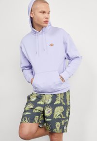 Sudadera de color púrpura claro con un bolsillo frontal y logo, combinada con pantalones cortos oscuros de diseño estampado que presentan motivos en verde y azul.