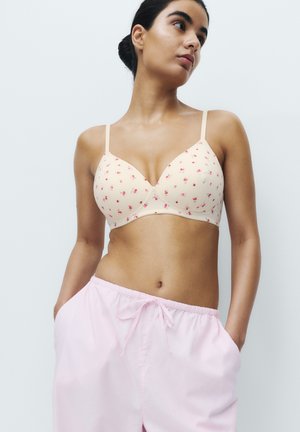 Donna con capelli scuri legati indietro che indossa un reggiseno crema con piccoli fiori rossi e pantaloni larghi rosa chiaro con coulisse, mani nelle tasche.