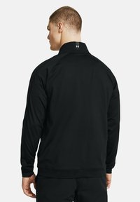 Chaqueta atlética negra con cuello alto, mangas largas, puños y dobladillo acanalados, que presenta un pequeño logo en la parte posterior del cuello. Textura suave.