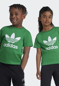 Zelené bavlněné trička s bílým logem Adidas a designem trojlístku, s krátkými rukávy a kulatými výstřihy, kombinované s černými kalhotami.