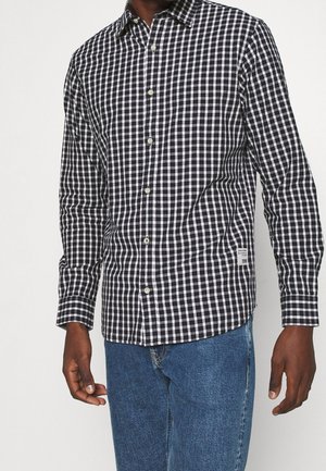 Chemise à carreaux noir et blanc en tissu léger, avec un col pointu et des manches longues. Petite étiquette logo sur le côté.