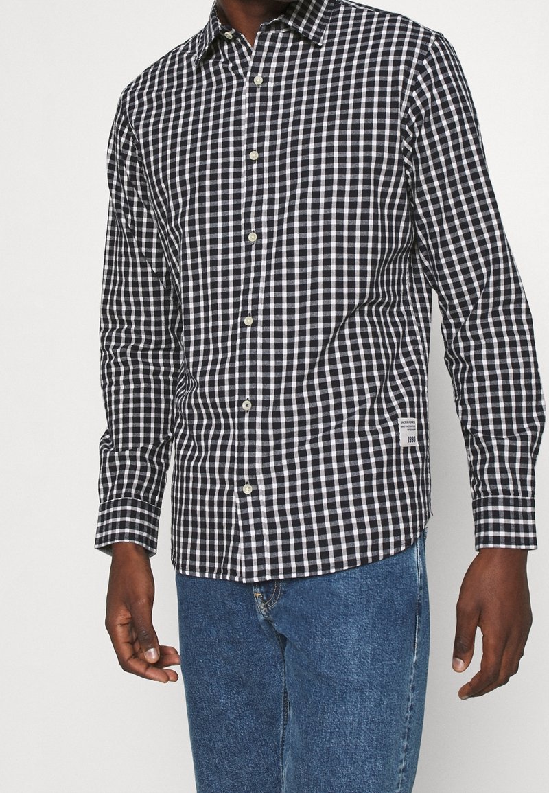 Camicia a quadri bianco e nero in un tessuto leggero, con colletto a punta e maniche lunghe. Piccola etichetta con logo sul lato.