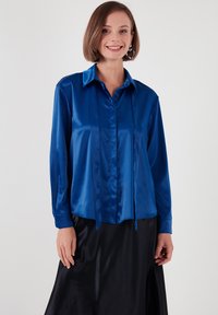 Blue satin button-up shirt with a collar, long sleeves, and a straight cut. Features vertical panels and a smooth texture.