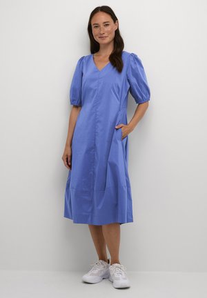 Vestito midi in cotone blu con maniche corte a palloncino, scollatura a V e tasche laterali; texture liscia e silhouette a trapezio. Indossato con sneakers bianche.