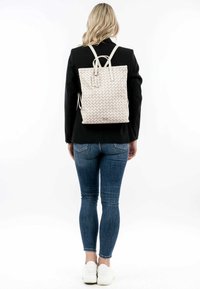 Sac à dos tissé blanc avec des bretelles réglables, en forme rectangulaire, à surface texturée et affichant un logo discret, porté par une personne en jean ajusté.