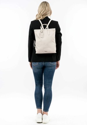 Zaino bianco intrecciato con spalline regolabili, forma rettangolare, superficie testurizzata e branding discreto, indossato da una persona in jeans aderenti.