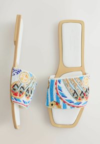 Paire de sandales plates blanches avec des brides larges décorées de perles colorées et complexes formant des motifs en bleu, rouge, jaune et noir.