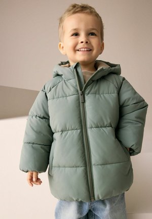 Kurtka puchowa w jasnozielonym kolorze, pikowana, z zapięciem na zamek i kapturem. Nosi na sobie beżowy sweter i niebieskie dżinsy.