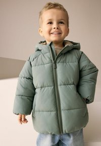 Green padded jacket with a zip closure and hood, featuring horizontal stitching and a soft lining, paired with denim jeans.