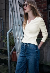 Lichtgeel henley-shirt met lange mouwen en knopen, gecombineerd met blauwe spijkerbroek met hoge taille. Op de achtergrond een metalen ladder en een bakstenen muur.