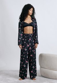 Ensemble de pyjama noir à motif floral comprenant un haut à manches longues boutonné et un pantalon large avec un motif de fleurs roses. Le tissu semble soyeux.