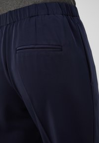 Pantalon bleu marine en tissu doux avec une texture lisse, présentant une poche arrière unique à ouverture étroite. Taille élastique pour plus de confort.