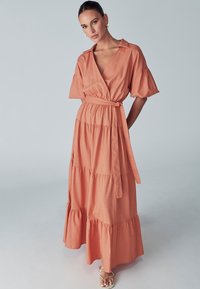 Maxi dress in coral, made of lightweight fabric. Features a wrap design, short sleeves, tiered skirt, and a tied belt at the waist.