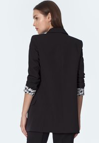 Kobieta w czarnym żakiecie z mankietami w kratkę houndstooth, widoczna z tyłu, na jednolitym jasnym tle.