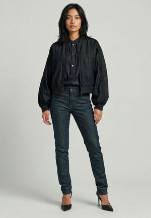 Mujer con cabello negro que lleva una chaqueta bomber negra, camisa azul oscuro con botones, vaqueros ajustados oscuros y tacones altos negros, de pie sobre un fondo liso.