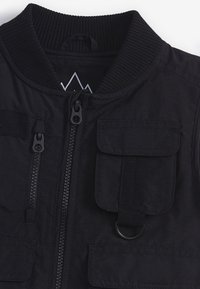Giacca bomber nera con colletto a costine, doppia zip, due tasche frontali e un anello per appenderla. Texture liscia con una finitura in tessuto resistente.