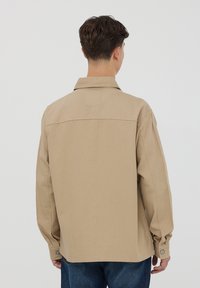 Camicia beige a maniche lunghe con colletto classico, polsini con bottoni e vestibilità rilassata. Realizzata in un tessuto testurizzato, con dettagli cuciti sul retro.
