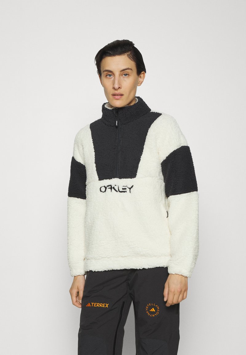 Person, der einen schwarz-weißen Fleece-Pullover mit "Oakley"-Text und schwarze Hosen mit orangefarbenen "Terrex"- und runden Logos trägt.