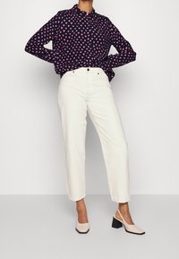 Blusa blu navy con motivo a cuori rosa, jeans bianchi a vita alta e scarpe con tacco a blocco beige. Tessuto liscio con vestibilità strutturata.