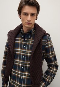 Camicia a quadri nei colori blu, verde e giallo, indossata sotto un gilet lavorato a maglia di colore marrone scuro con cappuccio.