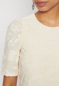 Beiges Texturkleid mit Puffärmel, floralem Muster und rundem Ausschnitt. Silbertöne Herzohrringe sichtbar.