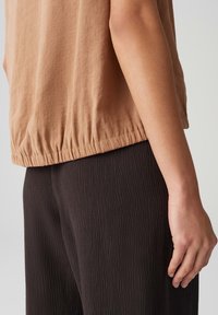 Top beige, testurizzato con orlo elastico abbinato a pantaloni neri a coste, con un primo piano del maniche e della mano su uno sfondo neutro.