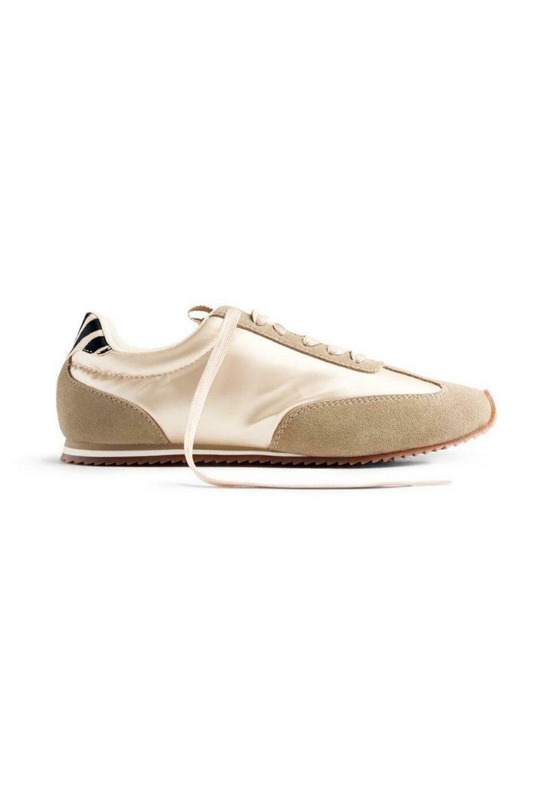 Zapatilla deportiva beige con una mezcla de materiales de satén y ante, diseño de cordones, puntera redondeada y suela de goma texturizada. Acento de talón a rayas.