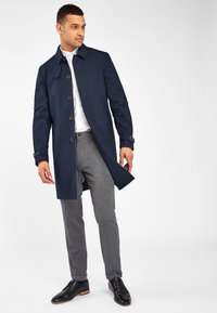 Trenca de color navy con cierre de botones, bolsillos laterales y puños ajustables, combinada con pantalones grises texturizados y zapatos de cuero negros.