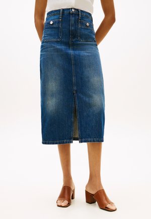 Denim-Midirock mit Frontschlitz, aufgesetzten Taschen, getragen mit weißem Oberteil und braunen, offenen Mules mit Absatz, vor weißem Hintergrund.