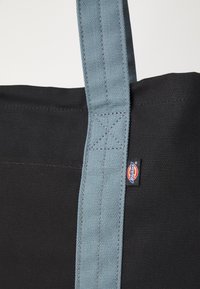 Μαύρη τσάντα ώμου με ανοιχτό μπλε λουρί. Διαθέτει ανθεκτικό ύφασμα, ορθογώνιο σχήμα και μια μικρή πινακίδα λογότυπου στο πλάι.