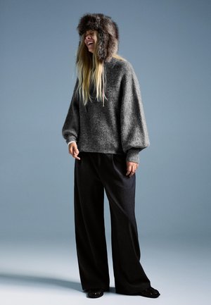 Pulover gri tricotat cu mâneci bufante, asortat cu pantaloni largi negri. O pălărie din blană artificială completează ținuta. Fundal simplu.