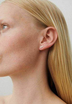 Lähivaade inimesest, kellel on blondid juuksed ja kes kannab vasakul kõrvakarval väikeseid, ümmargusi, kolmekordse täpiga naast-kõrvarõngaid neutraalse taustaga.