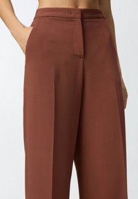 Pantalones de pierna ancha en una tela marrón óxido, con una textura suave, bolsillos frontales y un diseño entallado con una cinturilla plana en la parte delantera.