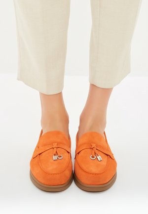 Mocassins en daim orange avec un bout rond, ornés d'un lacet décoratif et d'une garniture en argent, associés à un pantalon clair et sur mesure.