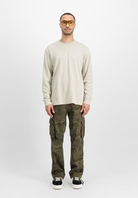 Beige Langarmshirt, Tarn-Cargo-Hose mit Seitentaschen, schwarze Sneaker mit weißen Schnürsenkeln und gelbe Sonnenbrille. Sauberes, lässiges Design.