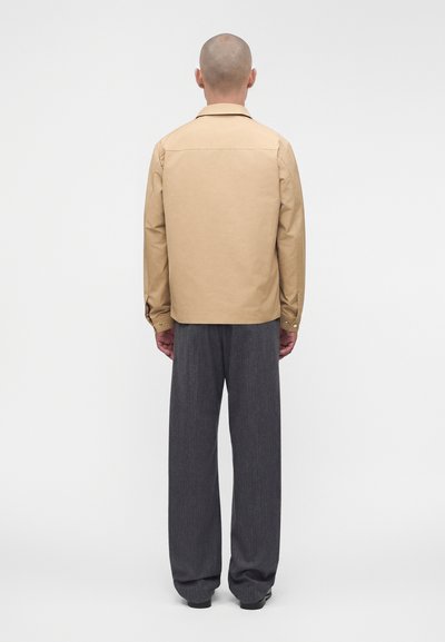 Homme à la tête rasée de dos, portant une chemise beige à manches longues et un pantalon ample gris foncé sur un fond clair uni.