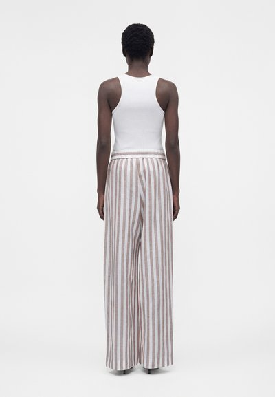 Άτομο που στέκεται με την πλάτη γυρισμένη φοράει λευκό αμάνικο τοπ και παντελόνι ψηλόμεσο, φαρδύ με κάθετες μπεζ και λευκές ρίγες.
