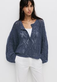 Gilet en tricot bleu foncé, présentant un motif en dentelle ouvert, un col rond et une fermeture à boutons. Manches longues et larges avec des poignets côtelés.