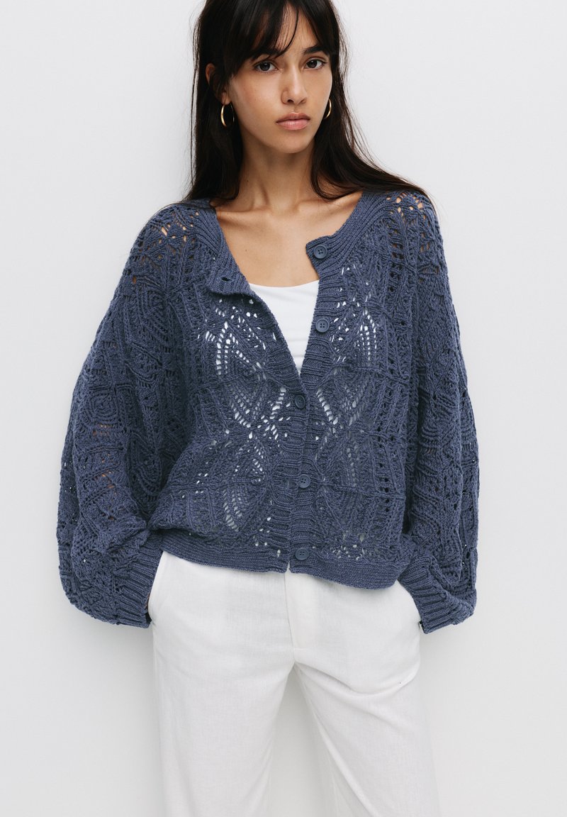 Gilet en tricot bleu foncé, présentant un motif en dentelle ouvert, un col rond et une fermeture à boutons. Manches longues et larges avec des poignets côtelés.