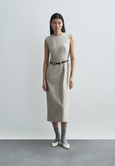 Μπεζ αμάνικο φόρεμα με υφασμάτινη υφή, μέχρι το γόνατο, μαζεμένο στη μέση με μια σκούρα ζώνη με χρυσές λεπτομέρειες. Συνδυασμένο με γκρι κάλτσες και λευκά παπούτσια.