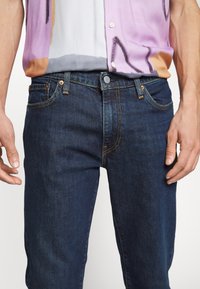 Homme portant un jean bleu foncé et une chemise multicolore boutonnée aux teintes violettes, blanches, grises et orange.