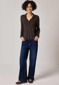 Frau steht mit Händen in den Taschen, trägt ein dunkelbraunes Hemd mit Knöpfen, weit geschnittene blaue Jeans und braune Schuhe, lächelt vor einem schlichten Hintergrund.