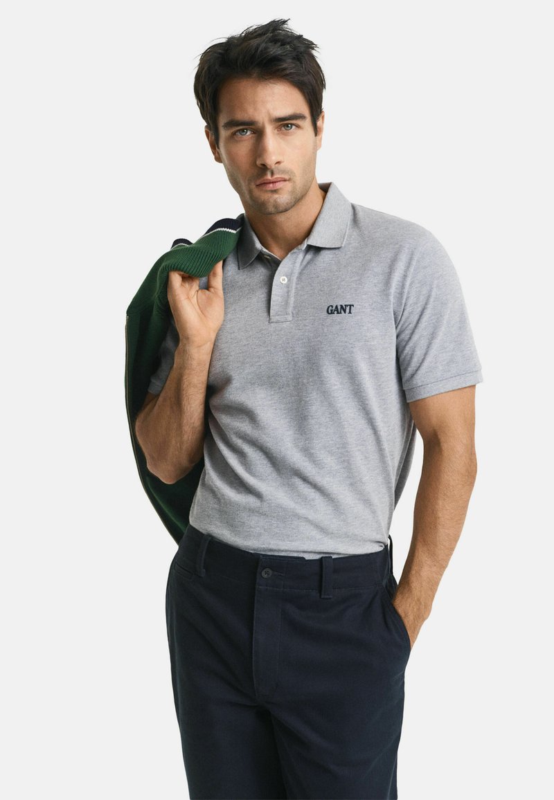 Mann in einem grauen GANT Poloshirt und dunklen Hosen, der eine grüne Jacke über der Schulter trägt, vor einem weißen Hintergrund.