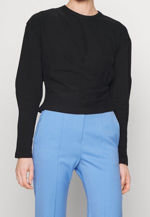 Osoba ubrana w czarną bluzkę z długim rękawem z bocznym zdobieniem i jasnoniebieskie, wysokiego stanu, dopasowane spodnie.