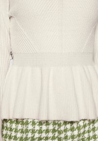 Krémově zbarvený pletený svetr s žebrovanou texturou a pasem s volánkem. Design má dlouhé rukávy a jemný vzhled.