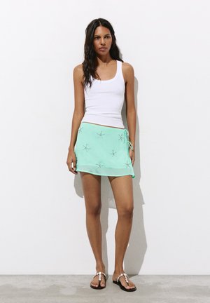 Femme portant un haut blanc sans manches, une mini-jupe vert menthe avec des motifs d'étoiles de mer, et des sandales noires ornées de perles blanches, debout contre un mur blanc.