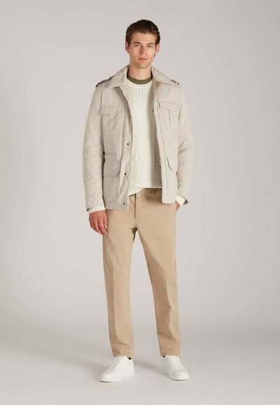 Jonge man in een beige jas over een witte kabelgebreide trui, khaki broek en witte sneakers, staand tegen een effen lichte achtergrond.