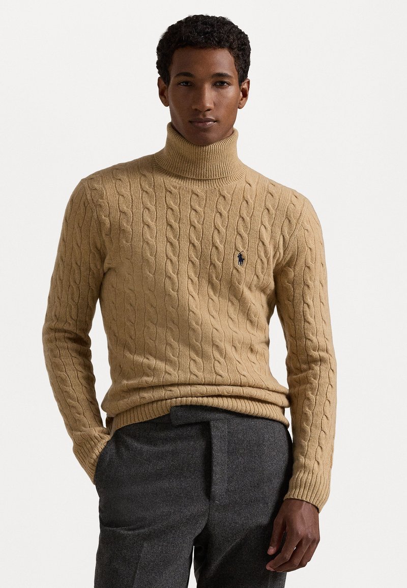 Tan kabelbrei coltrui met ribbels aan de mouwen en onderkant. Heeft een klein donker logo op de borst. Gekocht in combinatie met grijze op maat gemaakte broek.